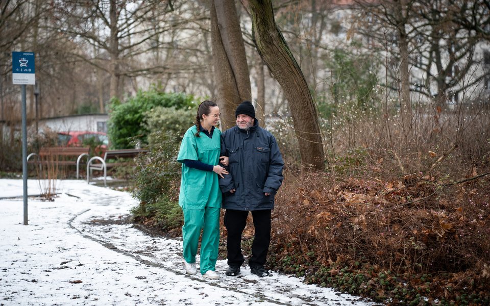Eine Pflegekraft in grüner Dienstkleidung geht untergehakt mit einem älteren Mann in Winterjacke einen verschneiten Parkweg entlang.