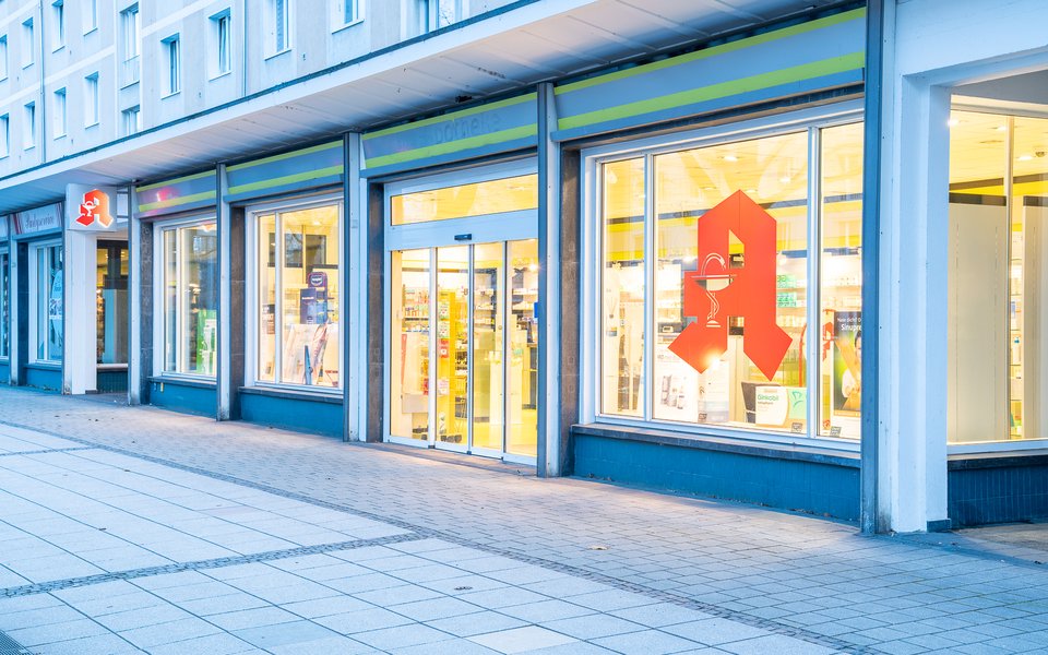 Die beleuchteten Schaufenster einer Apotheke mit rotem Apotheken-A in der Abenddämmerung, davor ein leerer Gehweg.